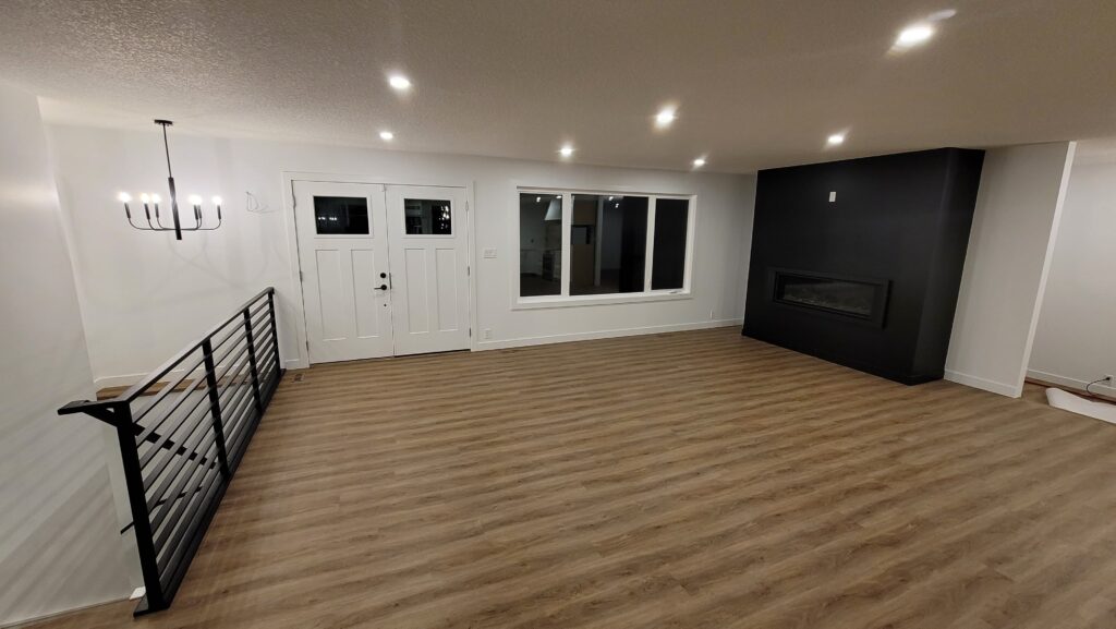 Open living room and entry after a whole home renovation in Edmonton, with a black linear fireplace, vinyl plank flooring, pot lights, and a black stair railing