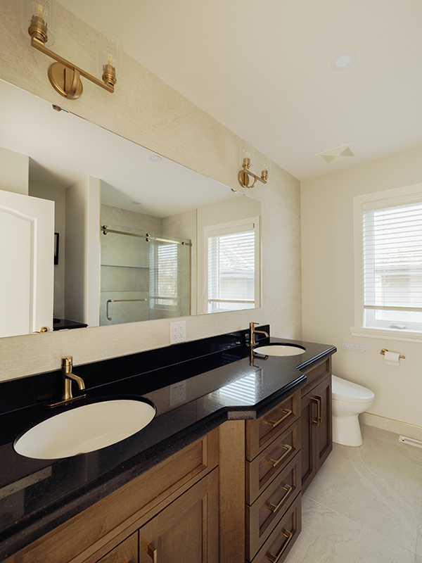 Double vanity with black granite countertop, dual undermount sinks, brass faucets, and brass sconce lighting above a full-width frameless mirror.