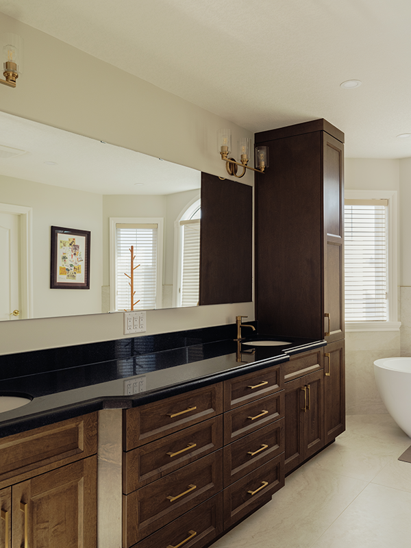 Dark maple vanity with black granite countertop, brass hardware, and a tall tower cabinet beside a freestanding tub in a renovated Edmonton bathroom.