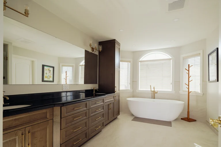 Wide view of an Edmonton bathroom renovation showing a dark maple double vanity, tower storage cabinet, brass fixtures, and a freestanding soaker tub beside arched windows.