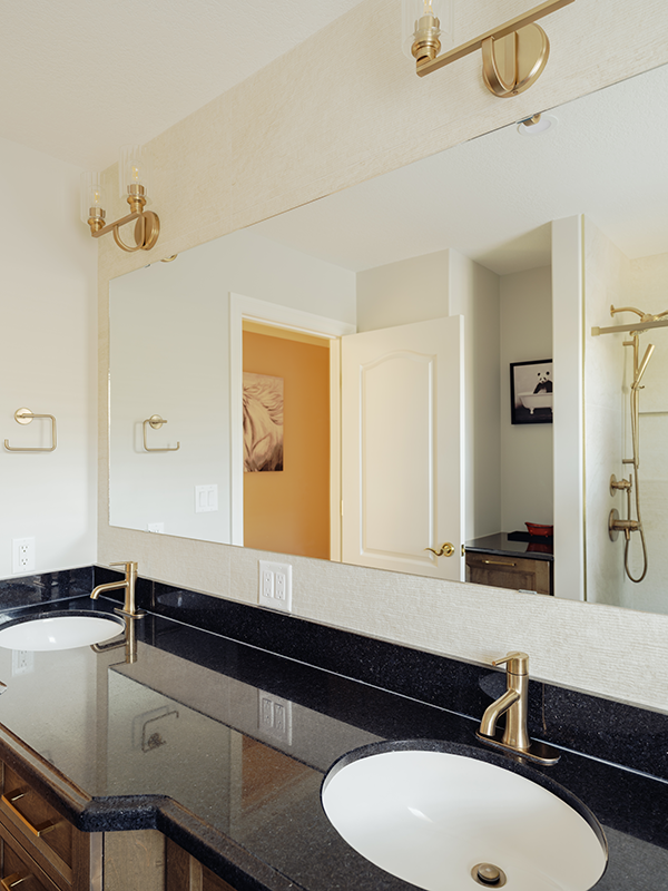 Black granite double vanity countertop with undermount sinks, brass faucets, and updated brass sconce lighting above a frameless mirror.