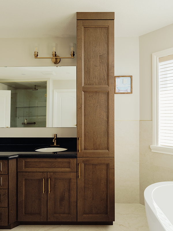 Tall maple tower storage cabinet beside a single-sink vanity with black granite countertop, brass hardware, and a freestanding tub in the background.