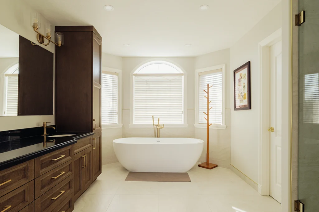 Renovated Edmonton primary bathroom with a white freestanding soaker tub, brass floor-mounted tub filler, dark maple vanity, and arched windows.