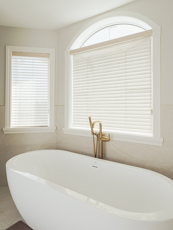 White freestanding soaker tub with a brass floor-mounted tub filler beneath an arched window with plantation blinds.