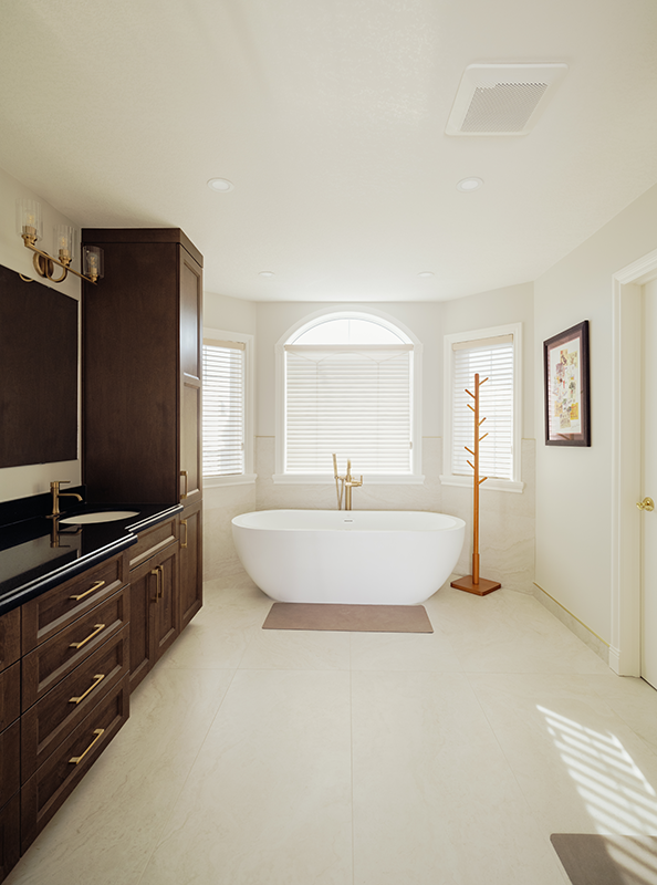 Primary bathroom renovation in Edmonton with a freestanding soaker tub, brass floor-mounted tub filler, dark maple vanity with tower storage, and large-format tile flooring.