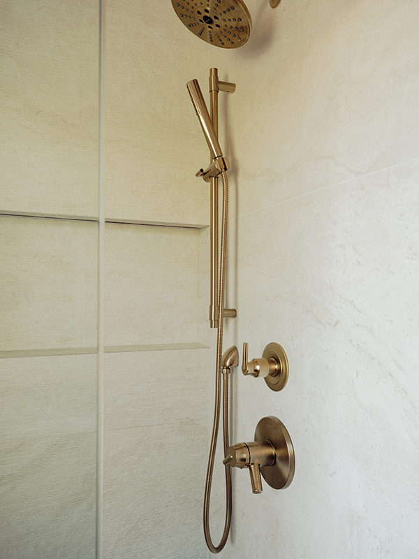 Detail shot of brass shower fixtures with slide bar, hand shower, and round control valves against marble-look tile.