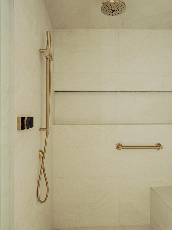 Close-up of brass shower fixtures including a rain head, slide bar, and grab bar against large-format marble tile.