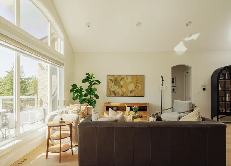 Open-concept living room with a vaulted ceiling, large picture window, brown leather sofa, and a framed floral art piece above a wood console.
