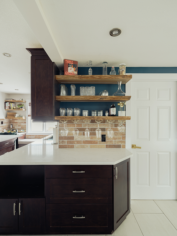 Wide view of an Edmonton kitchen renovation with reclaimed wood open shelving, brick tile backsplash, hanging stemware, navy accent wall, and a white quartz island.