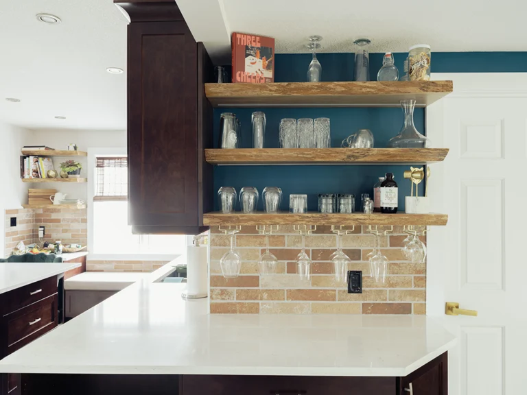 Reclaimed wood open shelving above a brick tile backsplash and stemware rack, beside dark espresso cabinets and a white quartz island.