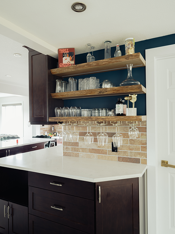 Kitchen bar area with reclaimed wood floating shelves, brick tile backsplash, stemware rack, and a navy accent wall behind dark espresso cabinetry.