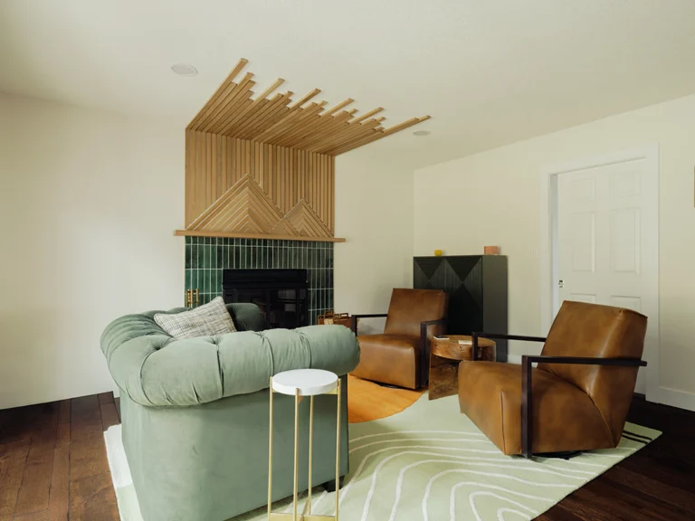 Close-up of a custom wood slat fireplace surround with chevron mantel detail and green tile surround, with leather chairs and a live-edge side table in the foreground.
