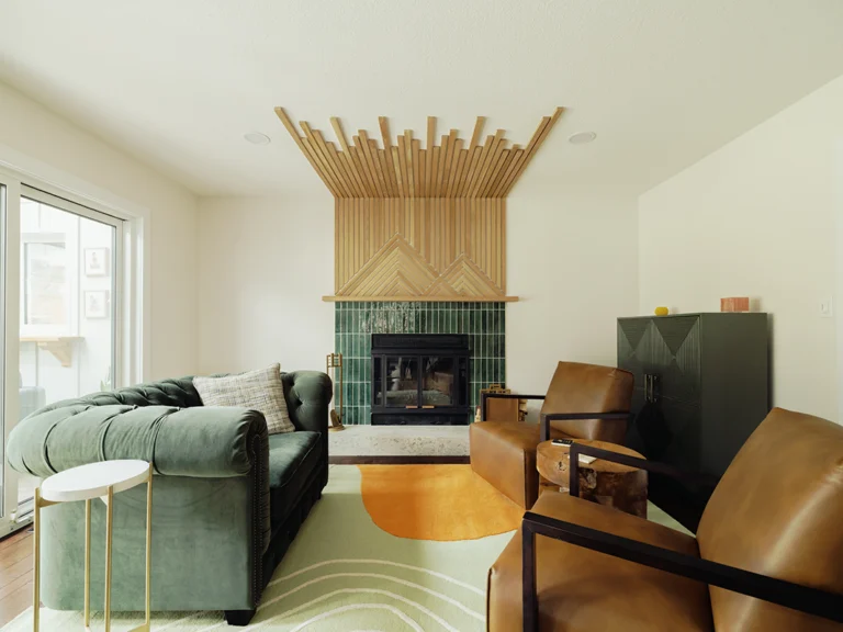 Living room renovation with a floor-to-ceiling wood slat fireplace surround, green tile firebox, green tufted sofa, leather chairs, and a patterned area rug.