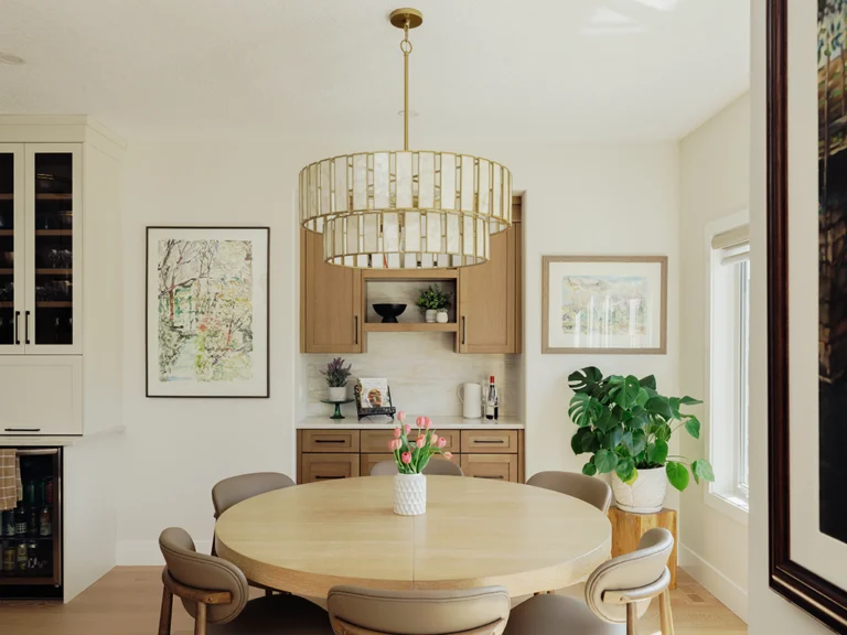 Wide-angle dining nook with a round oak table, upholstered chairs, brass drum chandelier, and a white oak coffee bar set between framed art.