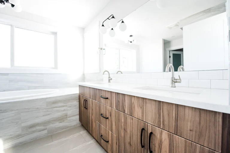 Wide view of primary ensuite in Edmonton home addition — walnut dual vanity with white quartz countertop, globe sconces, and soaker tub surround