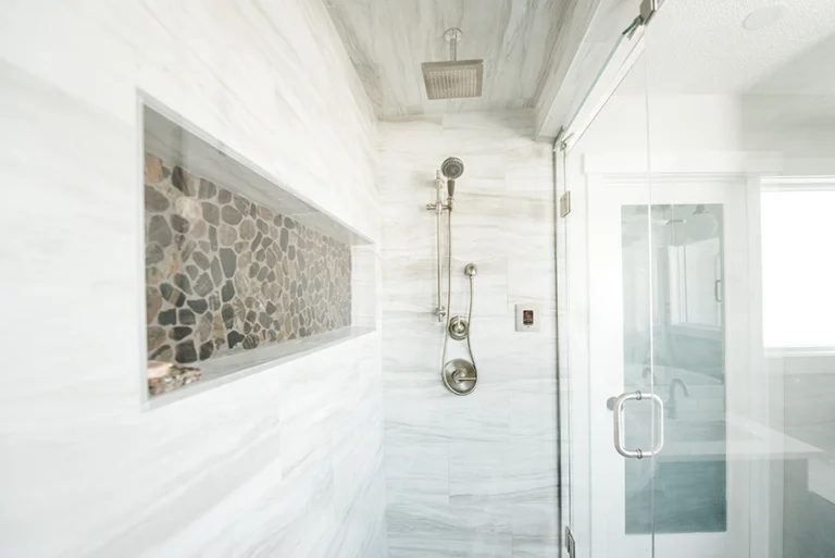 Interior of steam shower showing rain head, handheld fixture, river rock accent niche, and marble-look tile — Edmonton primary suite addition